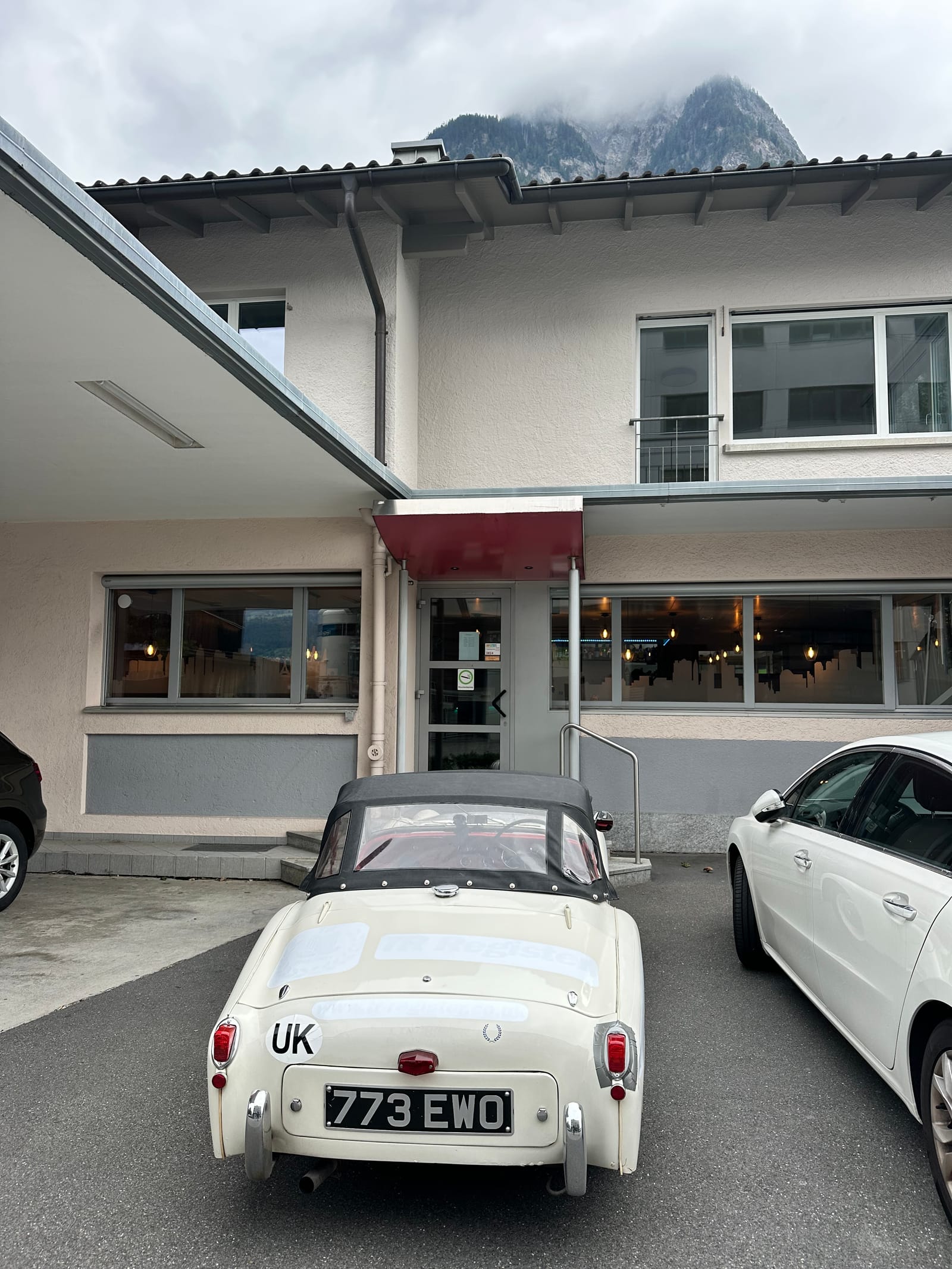 TS2 parked outside a cafe, hood up, a modern hatchback alongside and mountains in cloud above the roofline