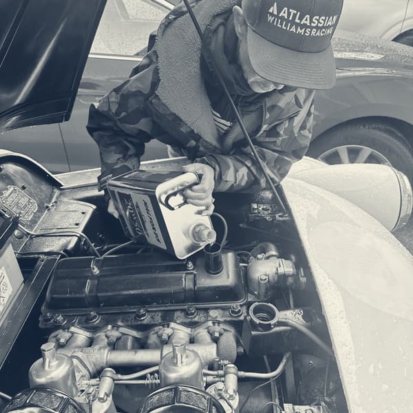 Evie leaning over TS2's open bonnet in a cap and dry robe, tipping oil from a green tin into the engine