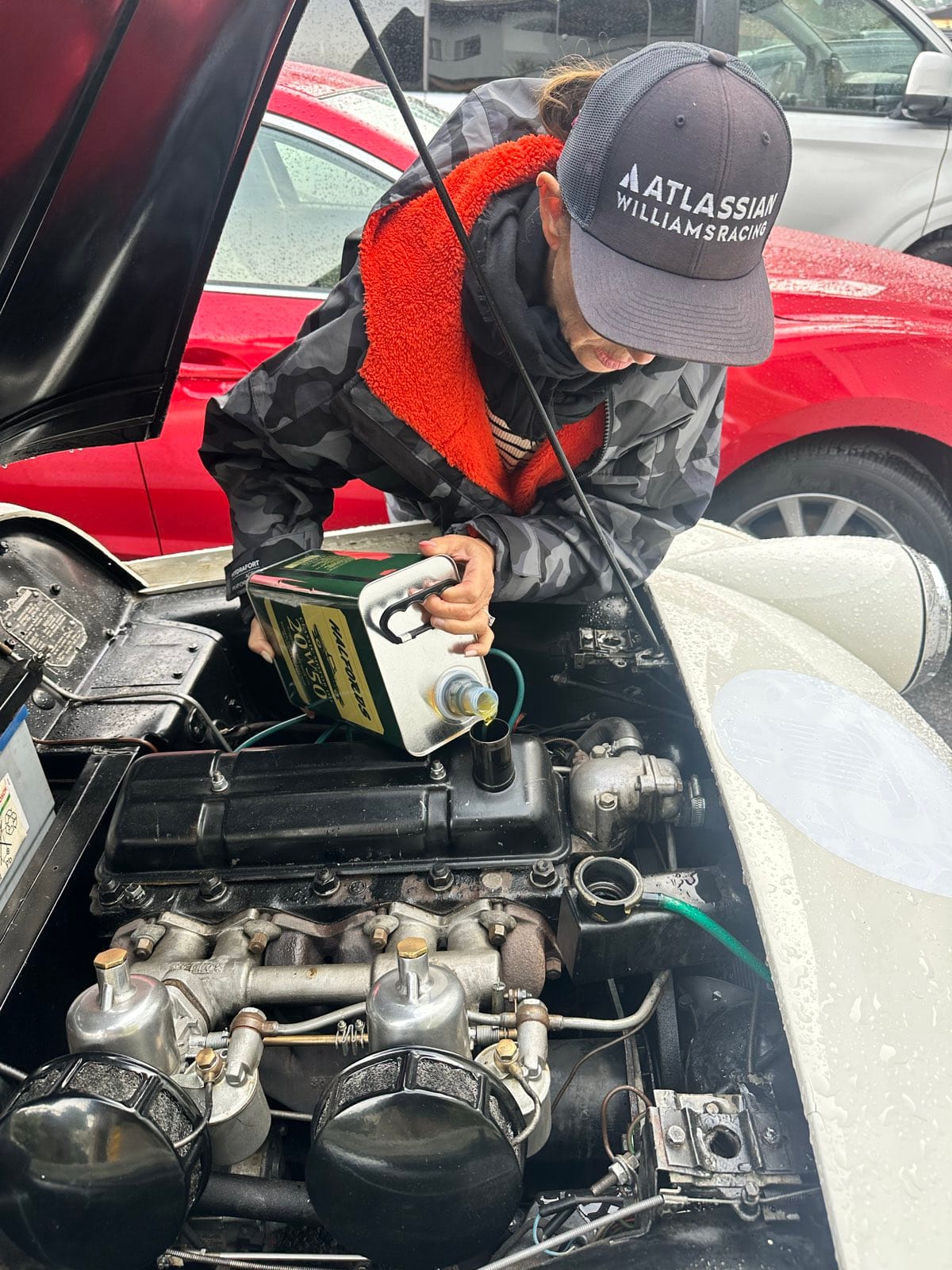 Evie leaning over TS2's open bonnet in a cap and dry robe, tipping oil from a green tin into the engine
