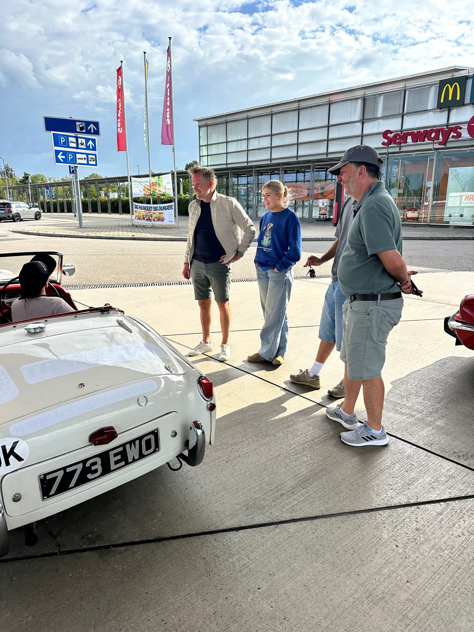 Three people and a girl in a blue jumper standing talking beside TS2 at a German service station, her number plate 773 EWO in the foreground