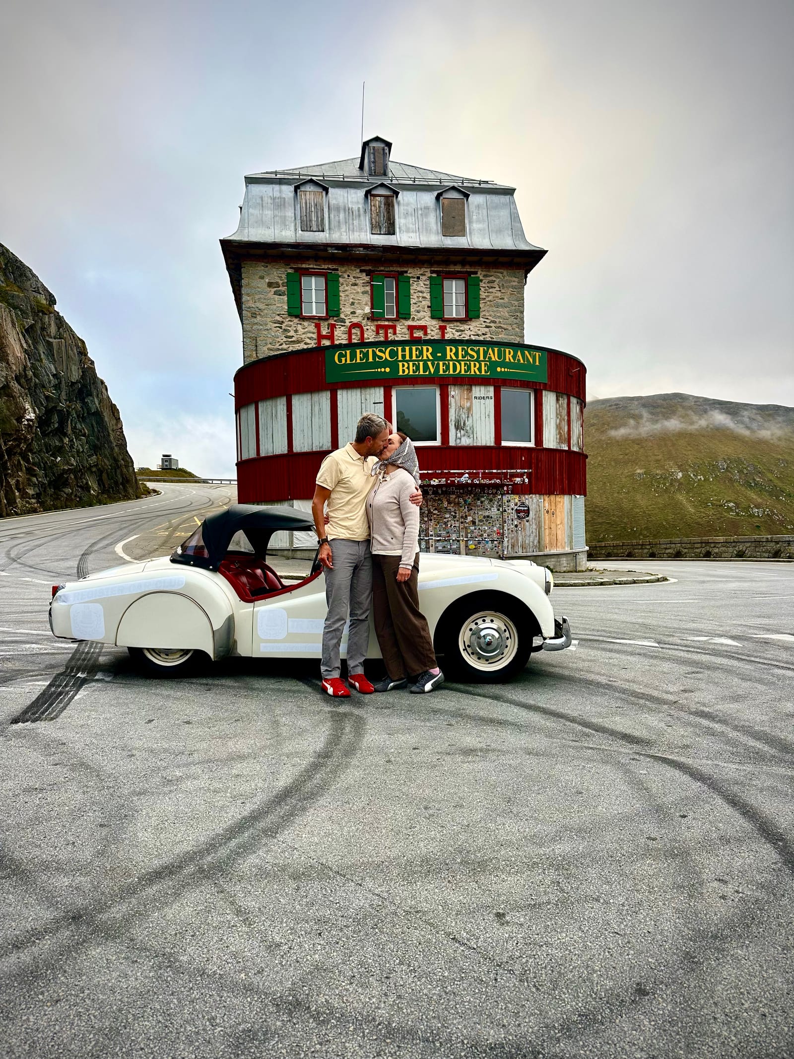 Evie and Andy kissing beside TS2's open door in front of the Hotel Belvédère, its red Gletscher Restaurant sign above them