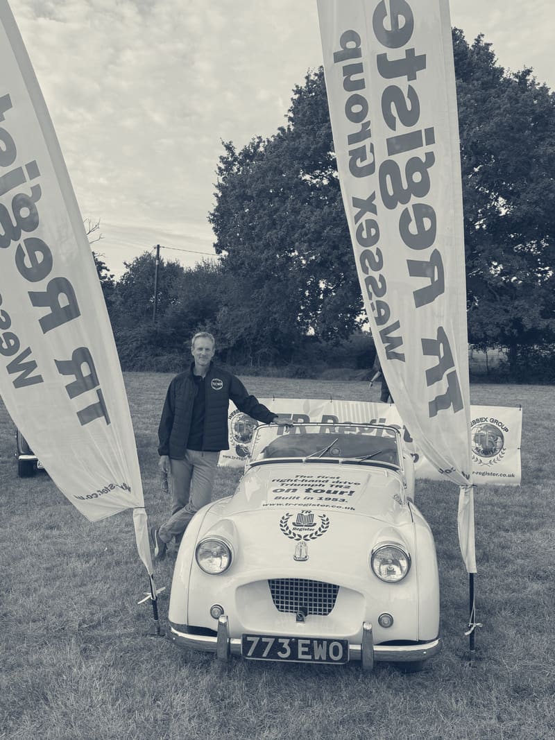 Andy standing beside TS2 in a field, a TR Register Wessex Group flag either side of the car
