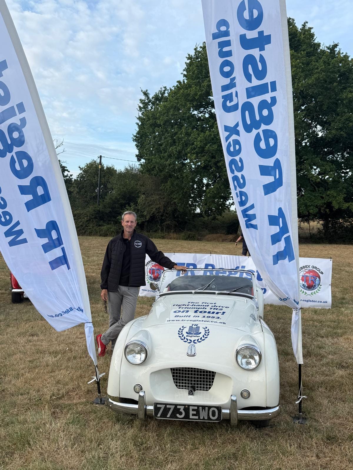 Andy standing beside TS2 in a field, a TR Register Wessex Group flag either side of the car