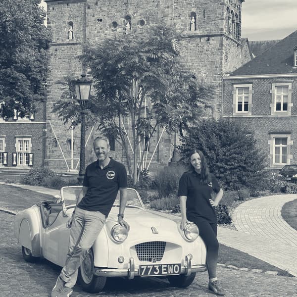 Andy and Evie in matching black TS2 polo shirts, leaning on TS2's headlamps on the cobbles in front of Rolduc Abbey, 773 EWO on the plate