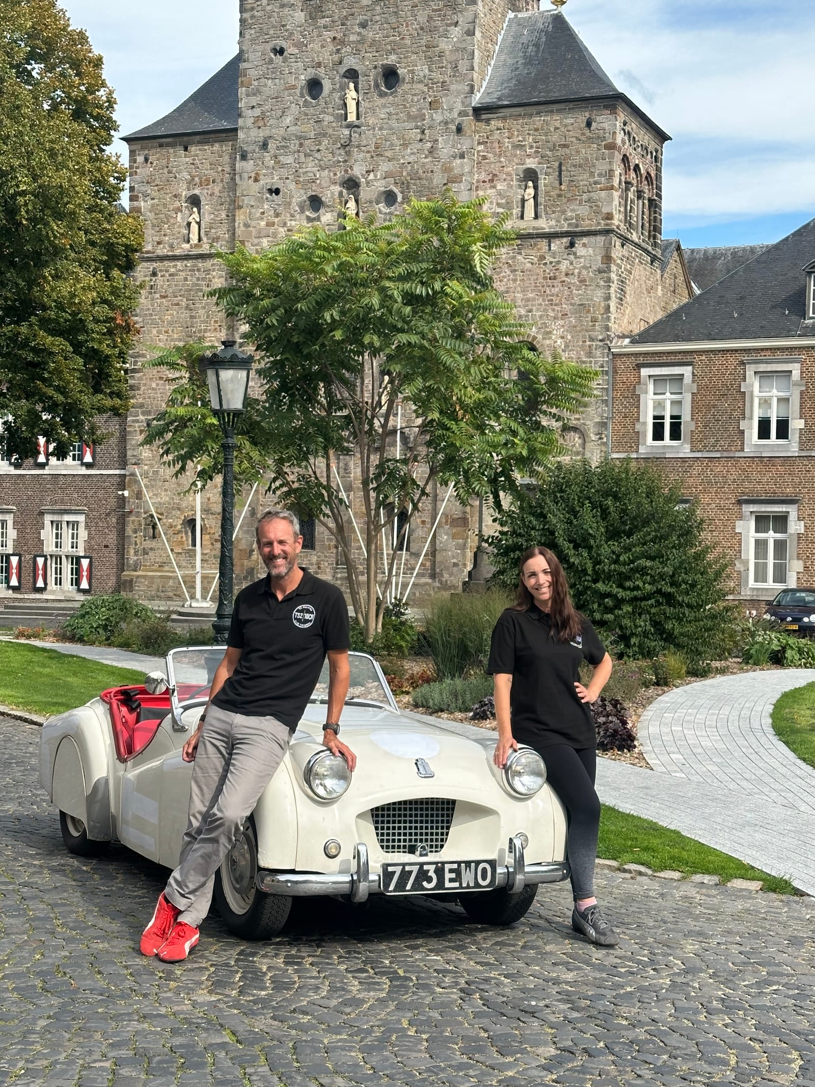 Andy and Evie in matching black TS2 polo shirts, leaning on TS2's headlamps on the cobbles in front of Rolduc Abbey, 773 EWO on the plate
