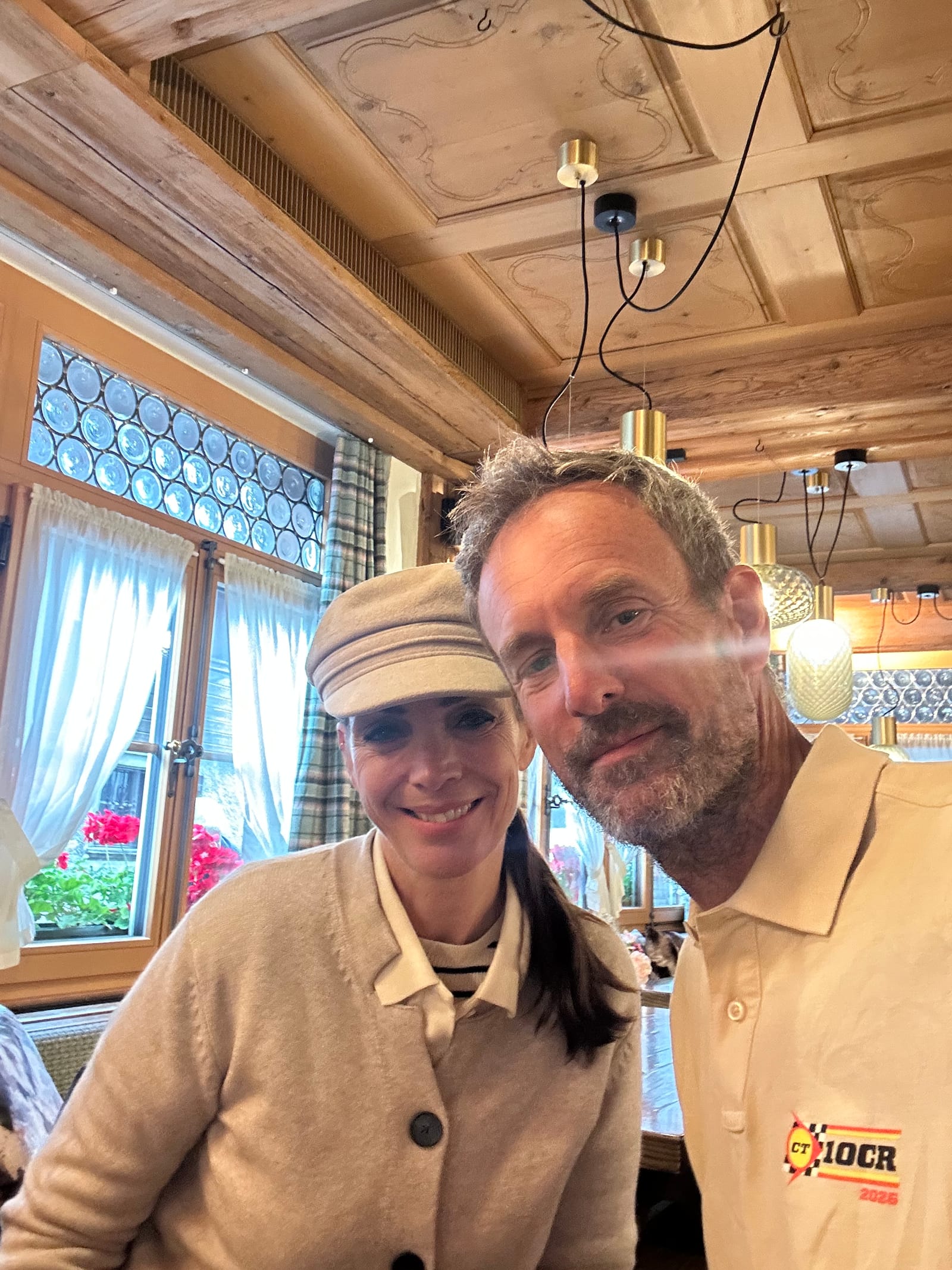 Evie and Andy cheek to cheek at the breakfast table, timber ceiling and leaded windows behind, Andy in his 10CR polo