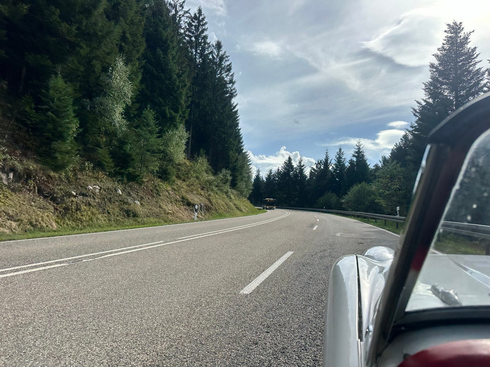 A Black Forest road curving away between tall pines, TS2's front wing and windscreen pillar in the near foreground
