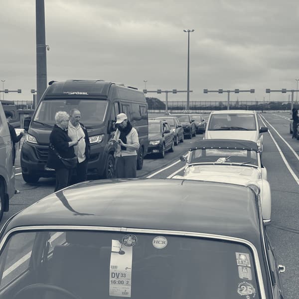 A boarding card reading DV33 16:00 taped inside the windscreen of a dark Triumph in the marshalling lanes, crews standing about between the cars, TS2 in the line beyond