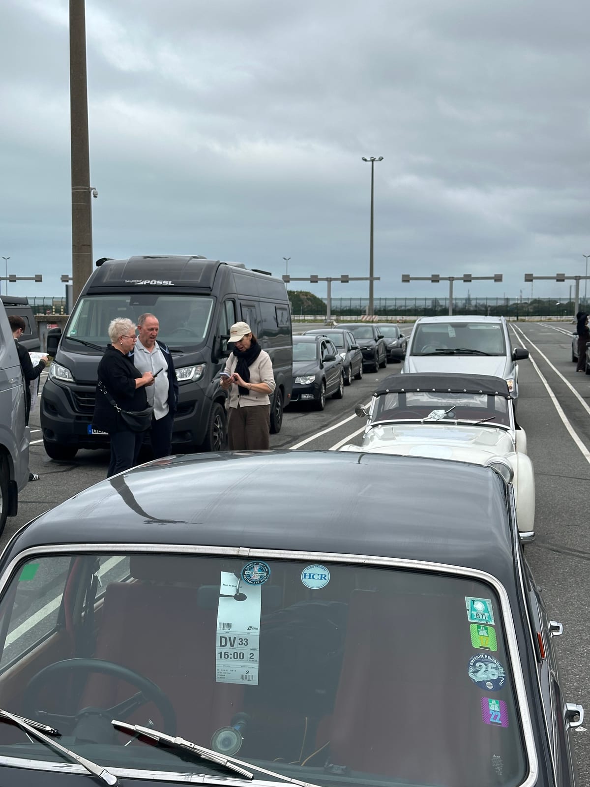 A boarding card reading DV33 16:00 taped inside the windscreen of a dark Triumph in the marshalling lanes, crews standing about between the cars, TS2 in the line beyond