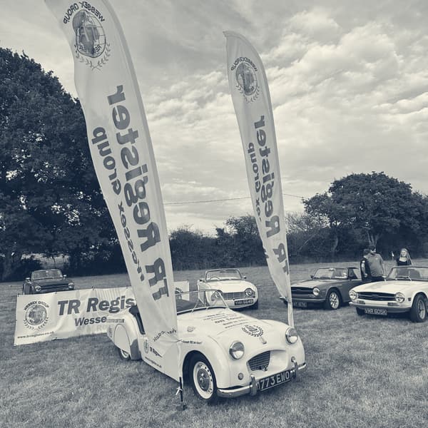 TS2 head on between two tall TR Register Wessex Group flags, the club banner behind and a row of later TRs to one side