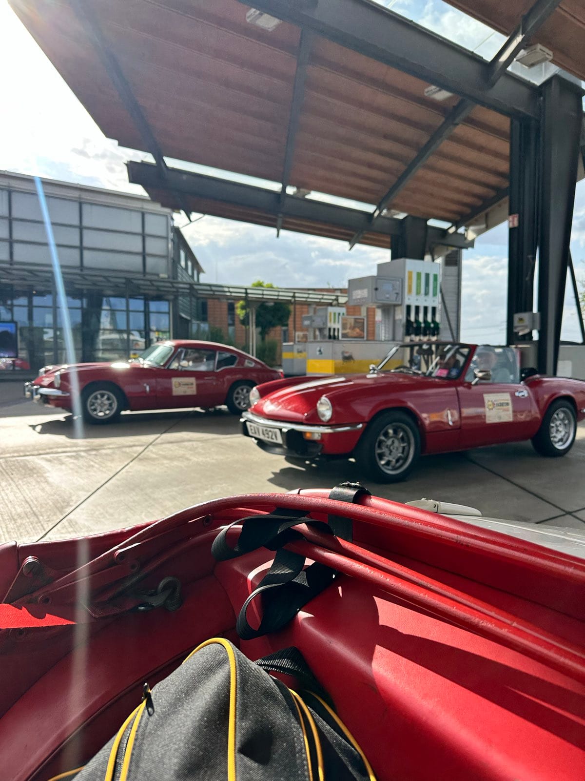Two red Triumphs, a GT6 and a Spitfire, parked at the same services, seen over TS2's folded hood