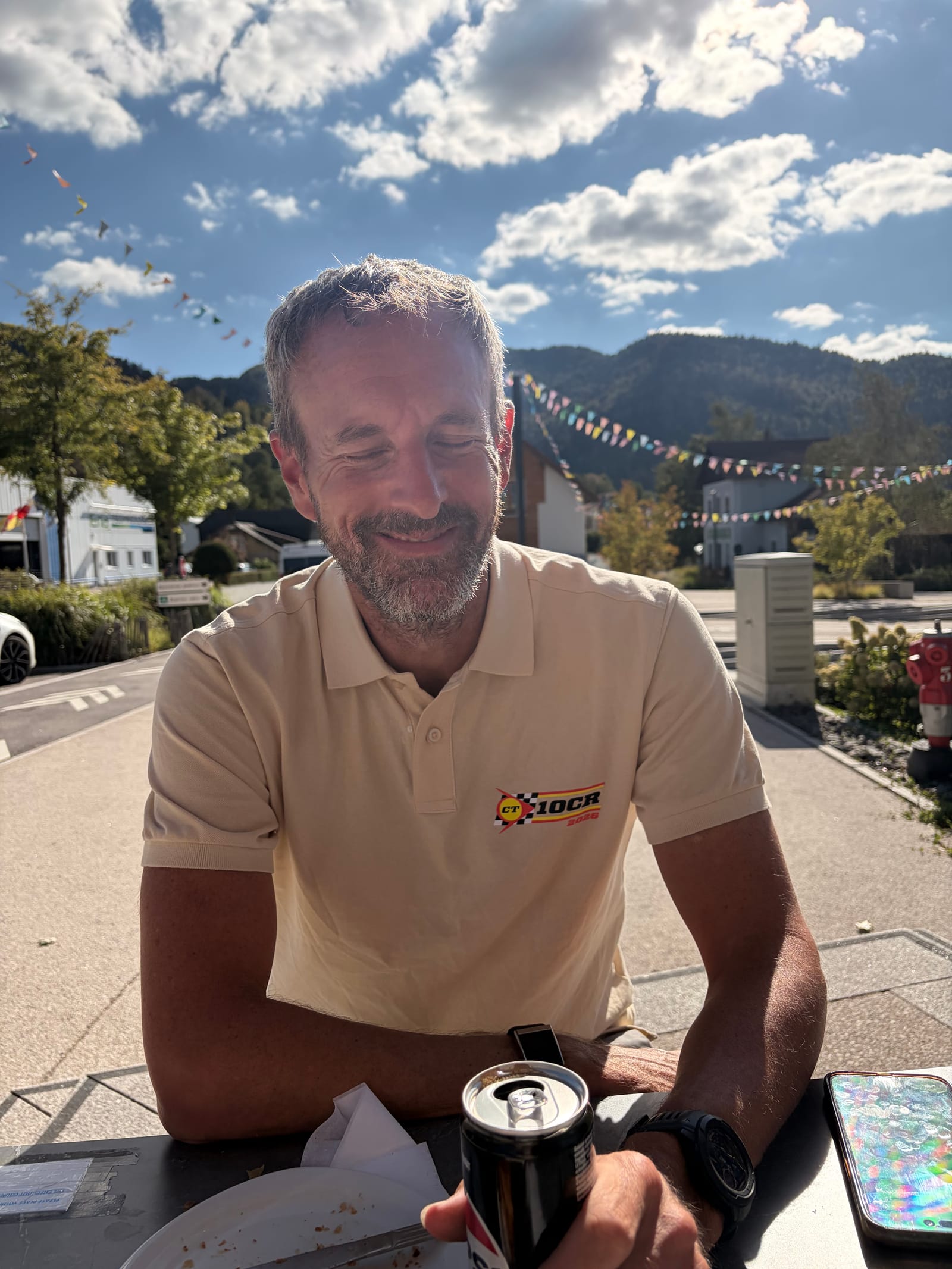 Andy at a table outside the patisserie with a steel travel cup, in his 10CR polo, the road book and a map underneath