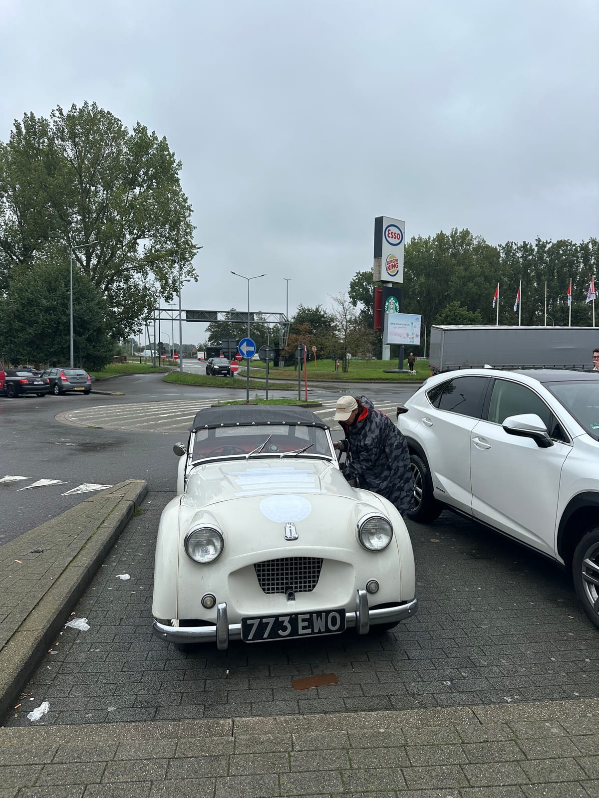 TS2 nose in at the kerb of an Esso services outside Antwerp, Evie in a hooded rain cape leaning into the cockpit, grey sky and a Burger King sign behind