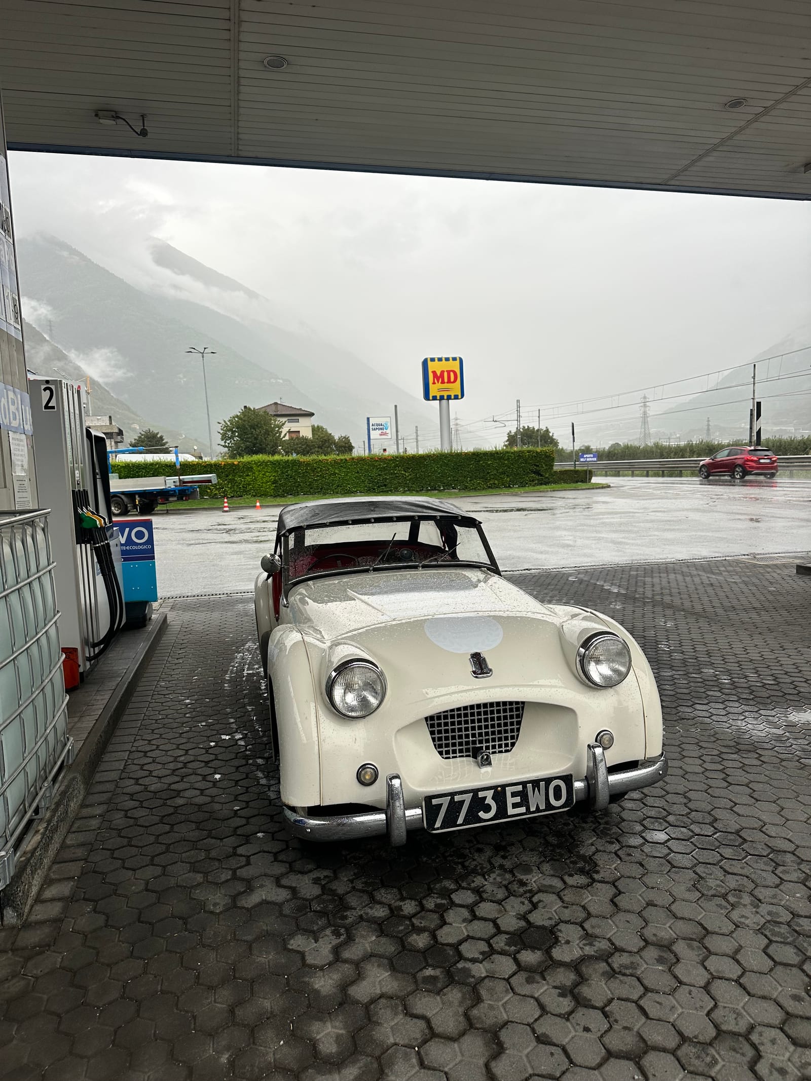 TS2 with her hood up under a filling station canopy, wet forecourt and a rain-grey valley behind