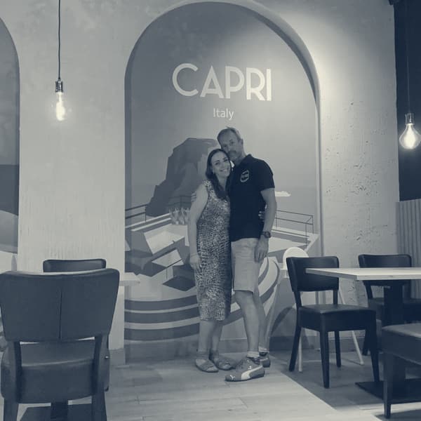 Evie and Andy arm in arm in an empty restaurant in front of a painted mural arch reading Capri, Italy