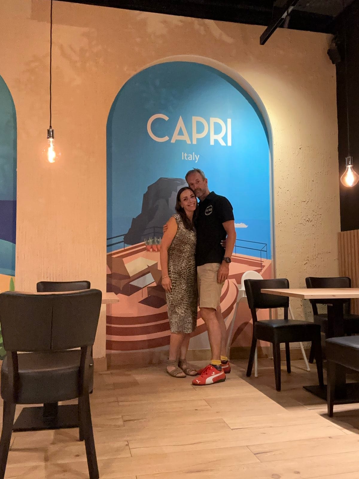 Evie and Andy arm in arm in an empty restaurant in front of a painted mural arch reading Capri, Italy