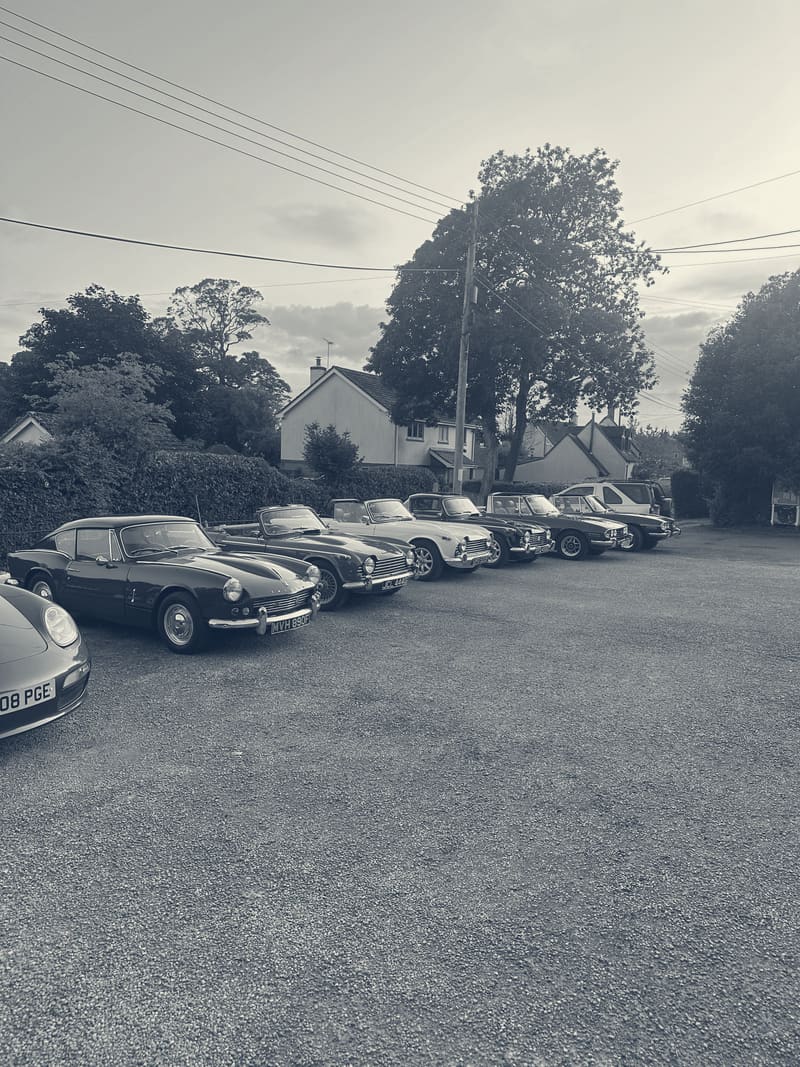 Triumphs lined up on gravel in low evening sun, a green GT6 nearest the camera, then a red TR4A, a white TR4 and a row of Stags.