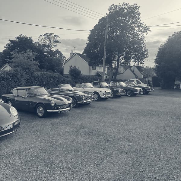 Triumphs lined up on gravel in low evening sun, a green GT6 nearest the camera, then a red TR4A, a white TR4 and a row of Stags.