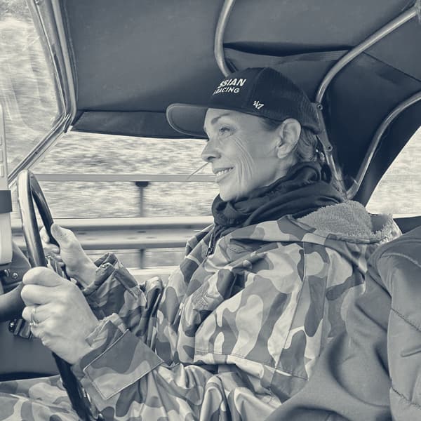 Evie at the wheel in her camouflage dry robe and cap, smiling, guardrail and trees behind