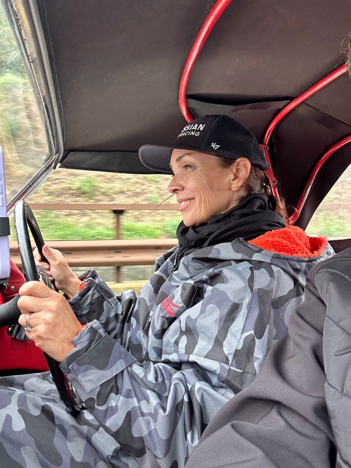 Evie at the wheel in her camouflage dry robe and cap, smiling, guardrail and trees behind