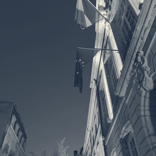 Looking up at the floodlit stone front of a building in Épinal, a French and an EU flag against a deep blue evening sky