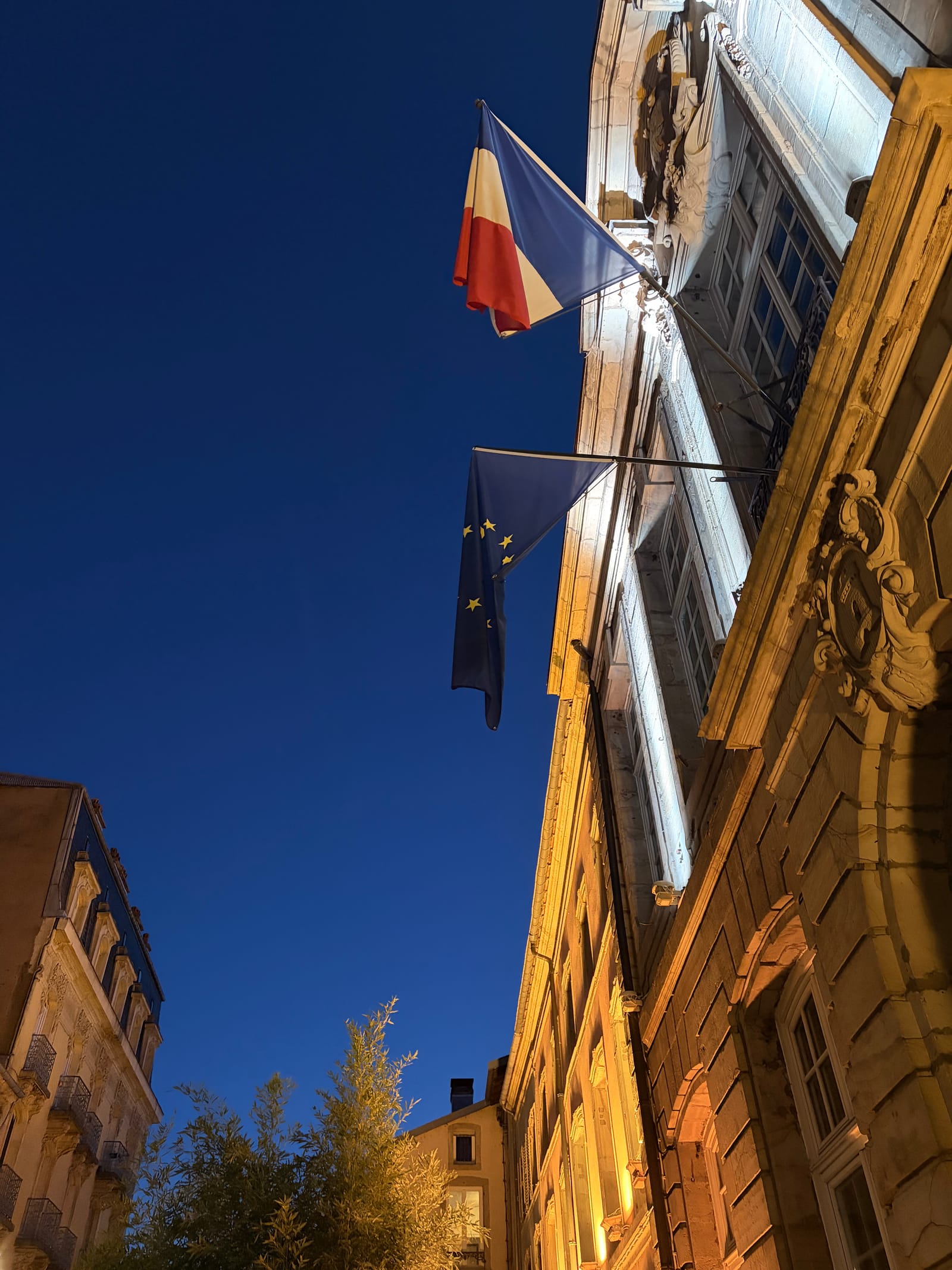 Looking up at the floodlit stone front of a building in Épinal, a French and an EU flag against a deep blue evening sky