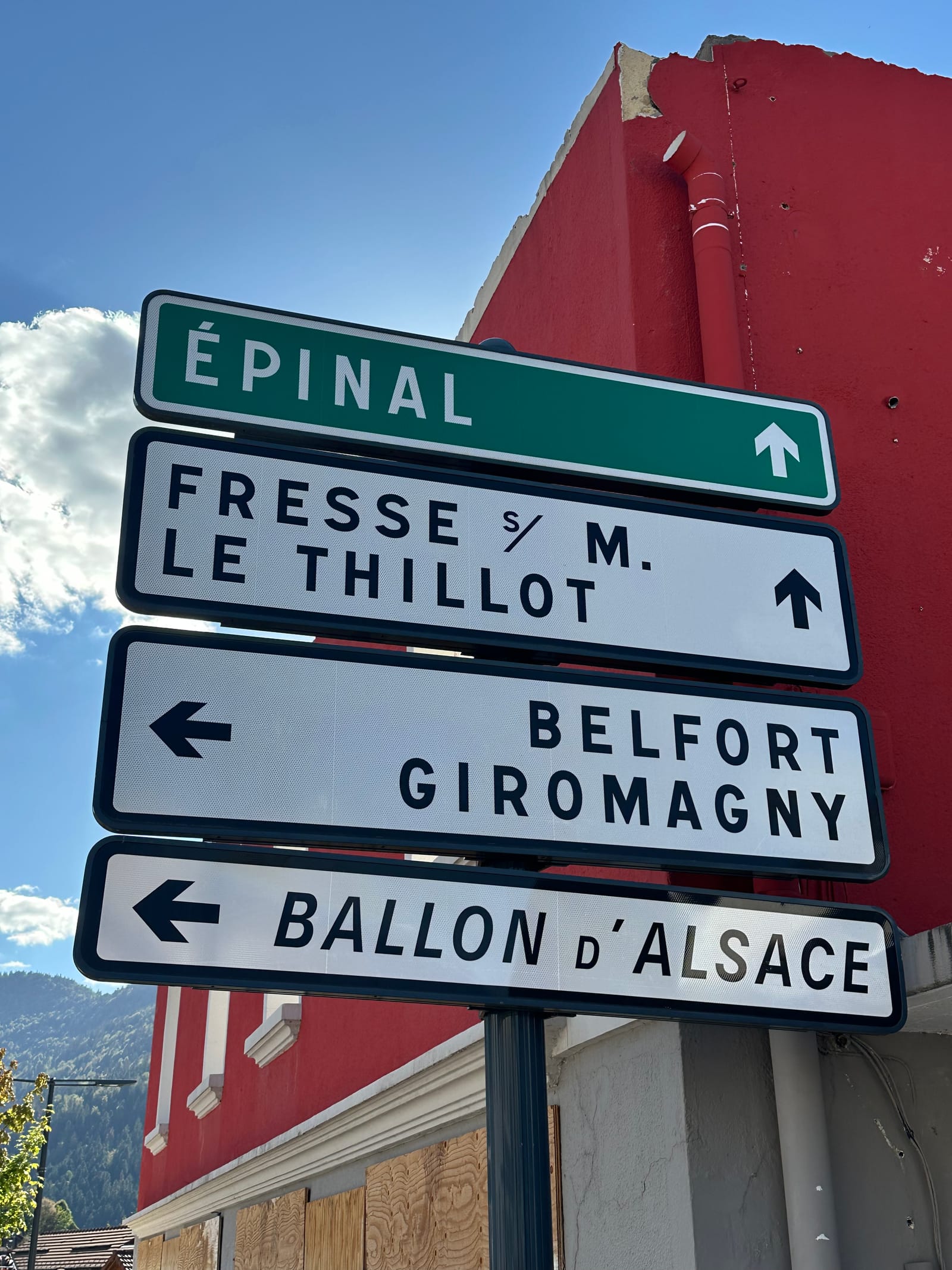 French road signs on a red-painted wall: Épinal straight on, Belfort and the Ballon d'Alsace to the left