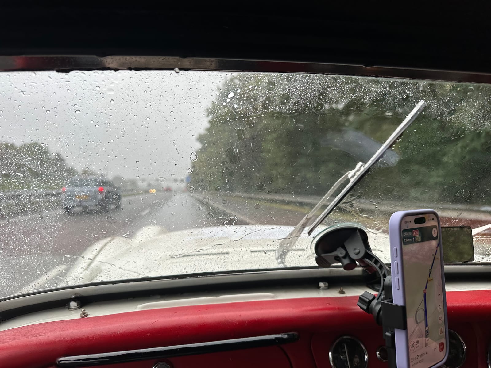 The view out of TS2's windscreen in the rain, wiper part way across, the phone on its mount showing the route, red dashboard below