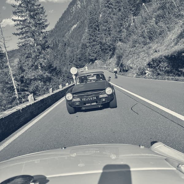 A dark blue TR6 ahead on a climbing road between spruces and rock