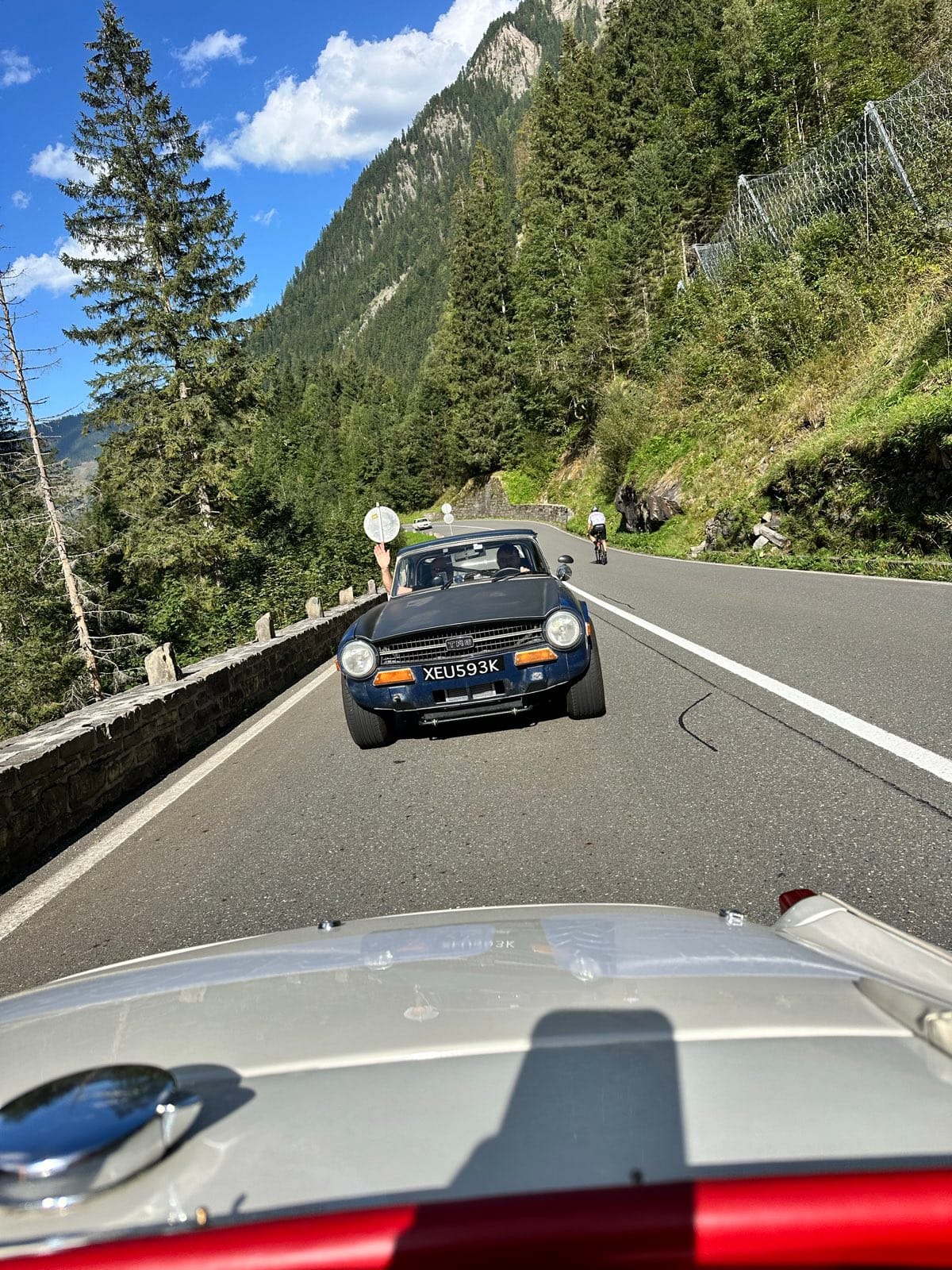 A dark blue TR6 ahead on a climbing road between spruces and rock