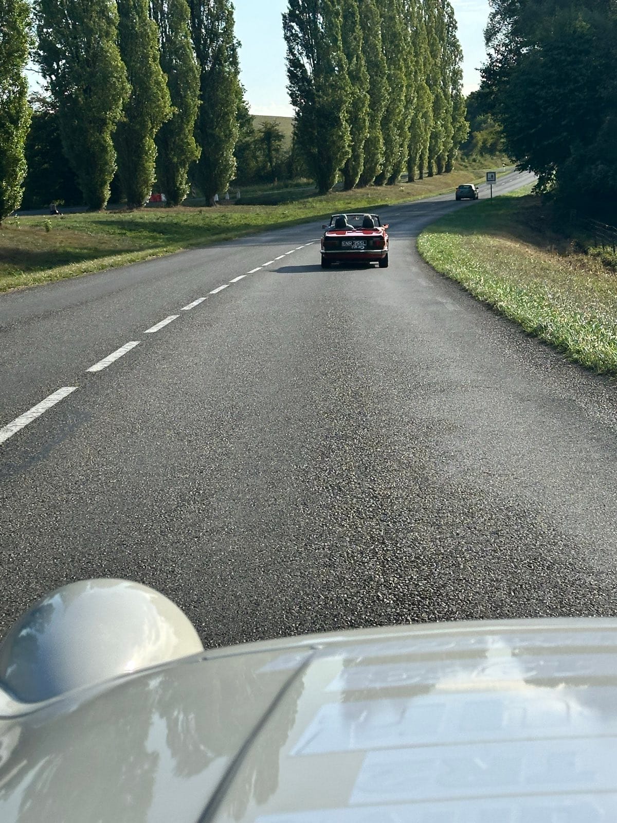 The view forward over TS2's bonnet down an avenue of poplars, a dark red TR6 ahead on the empty road