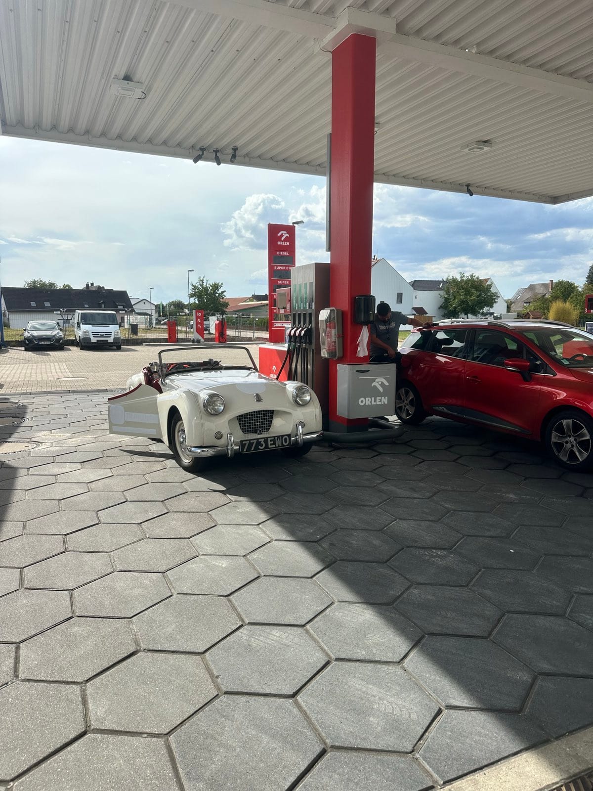 TS2 parked nose in under the canopy of a German filling station, hood down, a red hatchback at the next pump