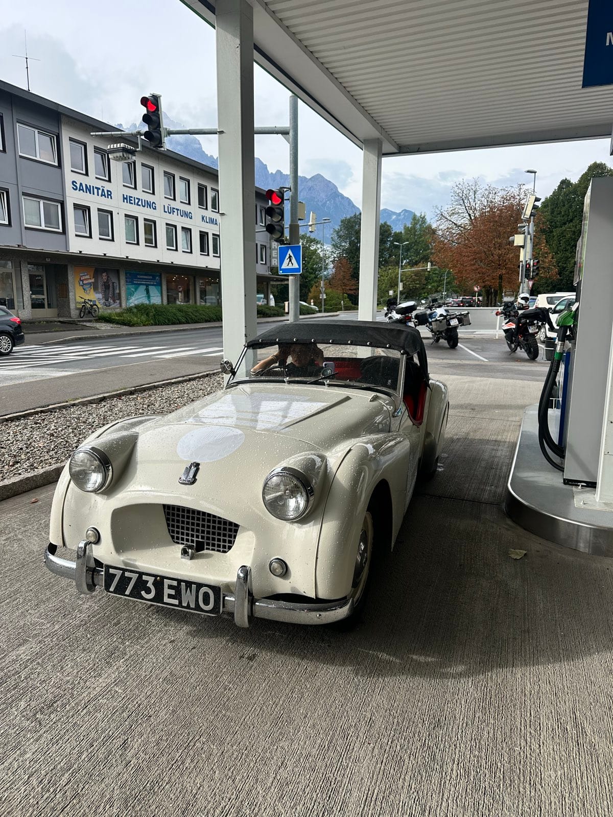 TS2 nose out under a filling station canopy, mountains and a row of motorcycles beyond