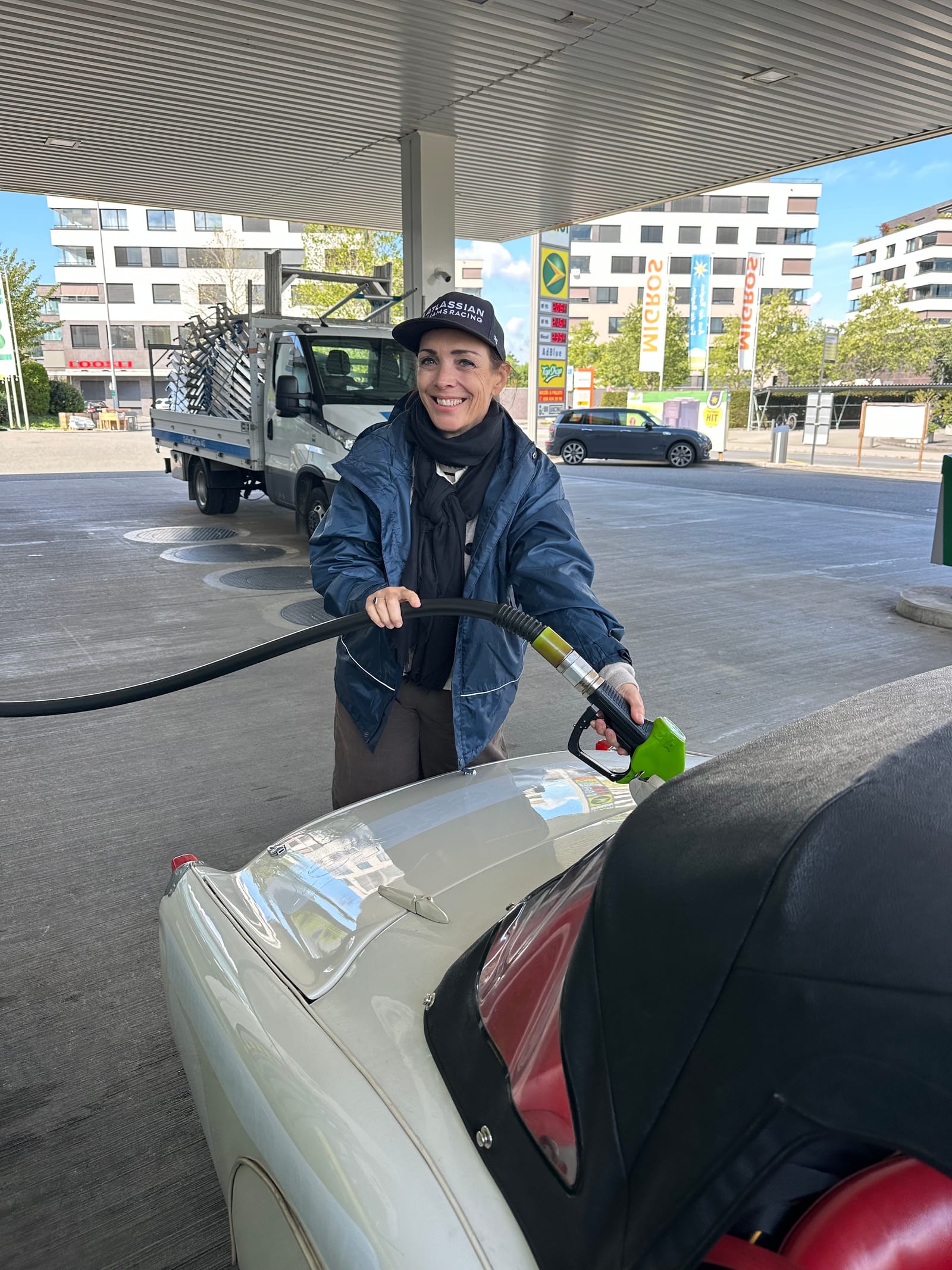 Evie in a cap and jacket holding the green pump nozzle to TS2's filler, a modern forecourt behind her