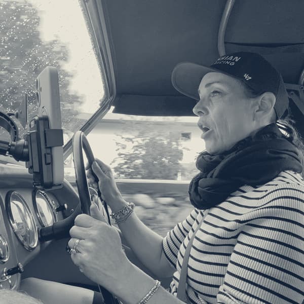Evie at the wheel under TS2's raised hood, mid sentence, hedges blurring past the windscreen