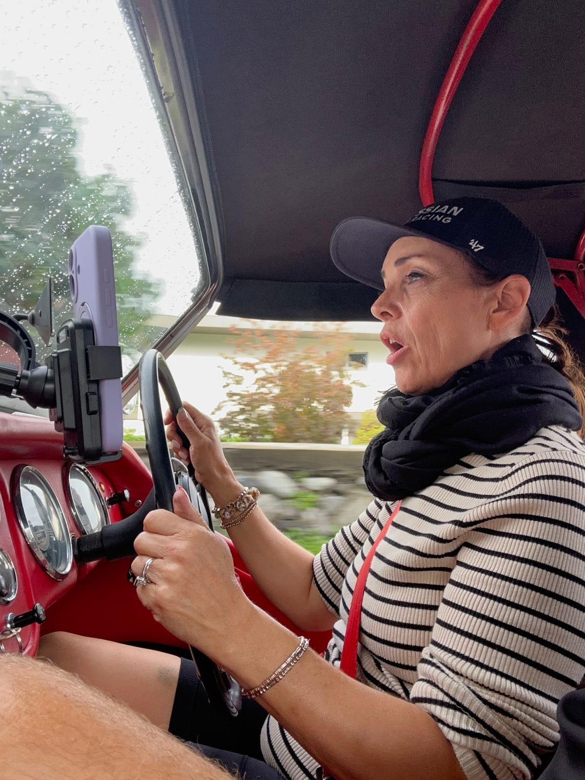 Evie at the wheel under TS2's raised hood, mid sentence, hedges blurring past the windscreen