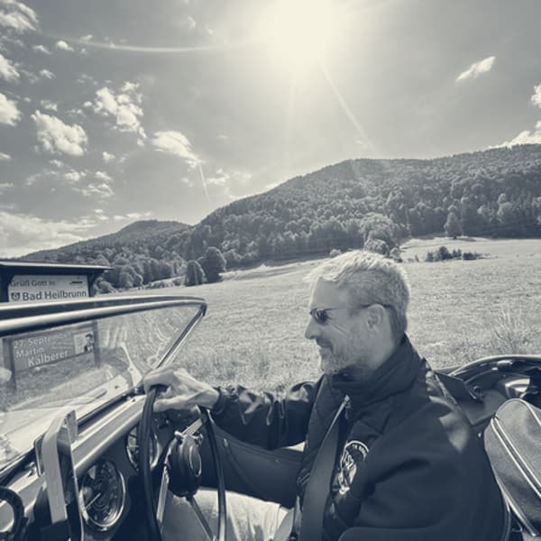 Andy at the wheel in sunglasses passing a roadside sign reading Gruess Gott in Bad Heilbrunn, wooded hill behind