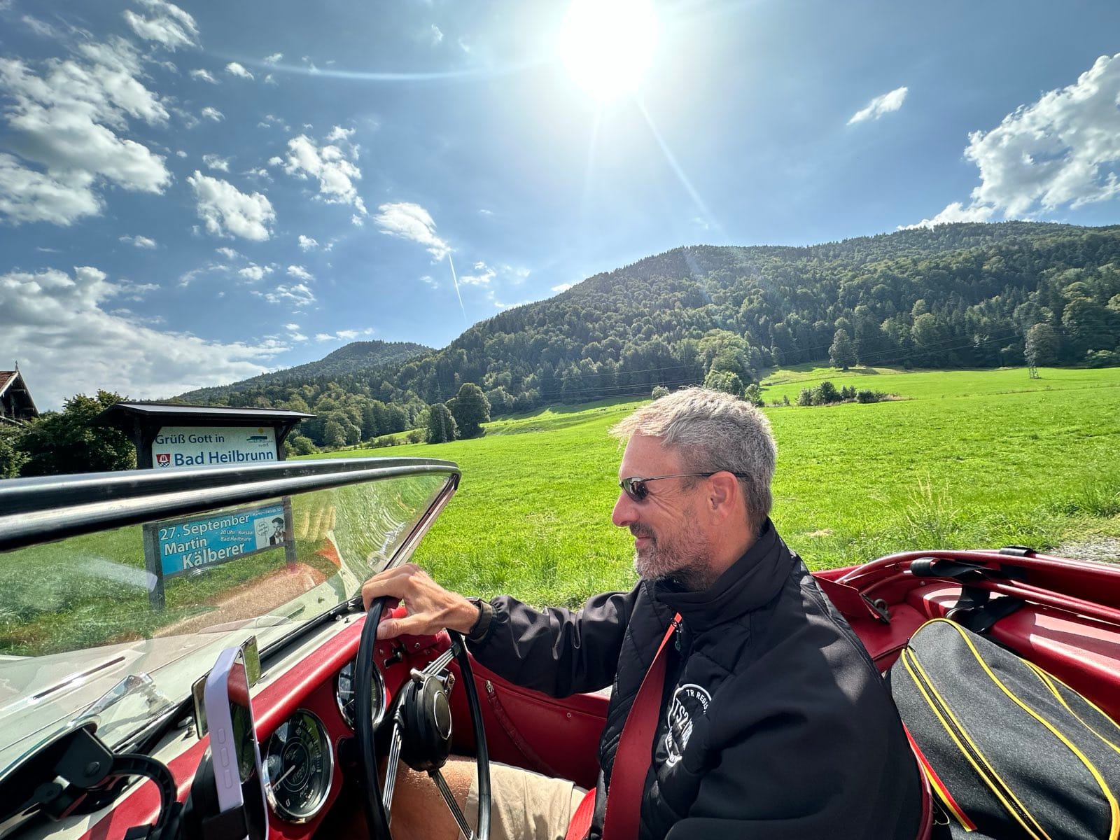 Andy at the wheel in sunglasses passing a roadside sign reading Gruess Gott in Bad Heilbrunn, wooded hill behind