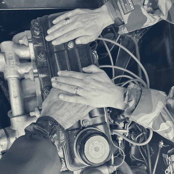 Two pairs of hands, one with a wedding ring, resting flat on the black rocker cover of TS2's engine