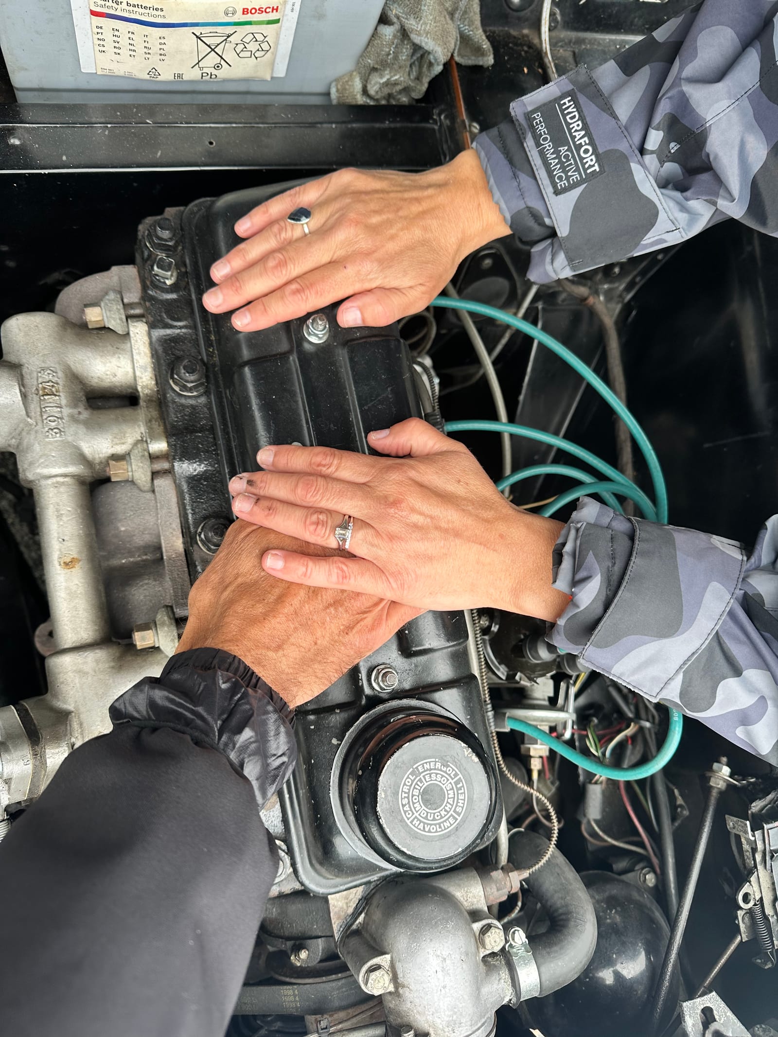 Two pairs of hands, one with a wedding ring, resting flat on the black rocker cover of TS2's engine