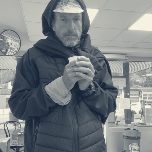 Andy in a hood and puffer jacket cradling a paper coffee cup with both hands, the pink cloth still on his wrist