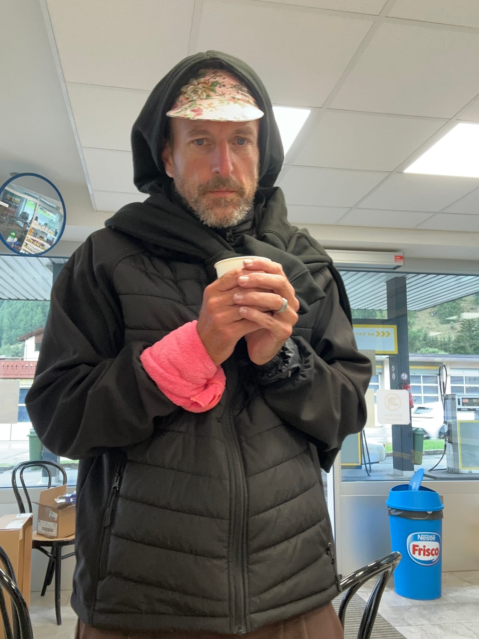 Andy in a hood and puffer jacket cradling a paper coffee cup with both hands, the pink cloth still on his wrist