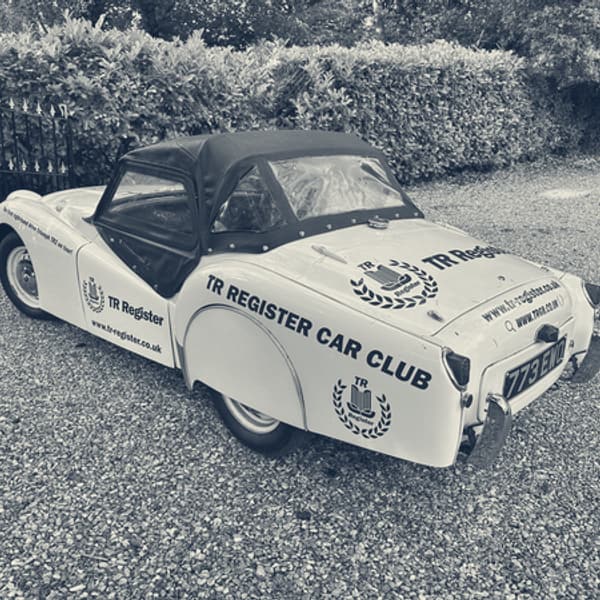 TS2 on the gravel drive from the rear three quarter, black hood up and sidescreens fitted, TR Register Car Club lettering along the rear wing