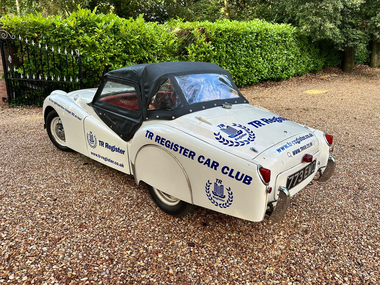 TS2 on the gravel drive from the rear three quarter, black hood up and sidescreens fitted, TR Register Car Club lettering along the rear wing