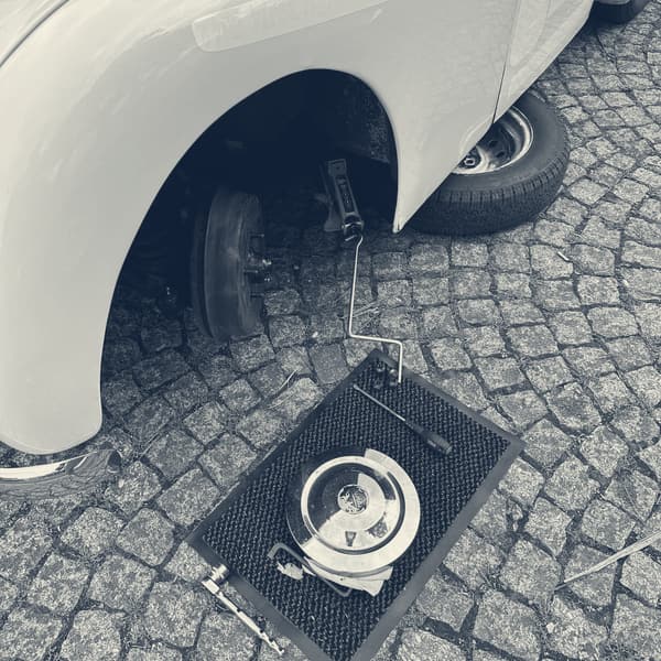 TS2 up on a trolley jack on the cobbles, front wheel off and the bare brake drum showing in the arch, the hubcap and a screwdriver laid out on the doormat