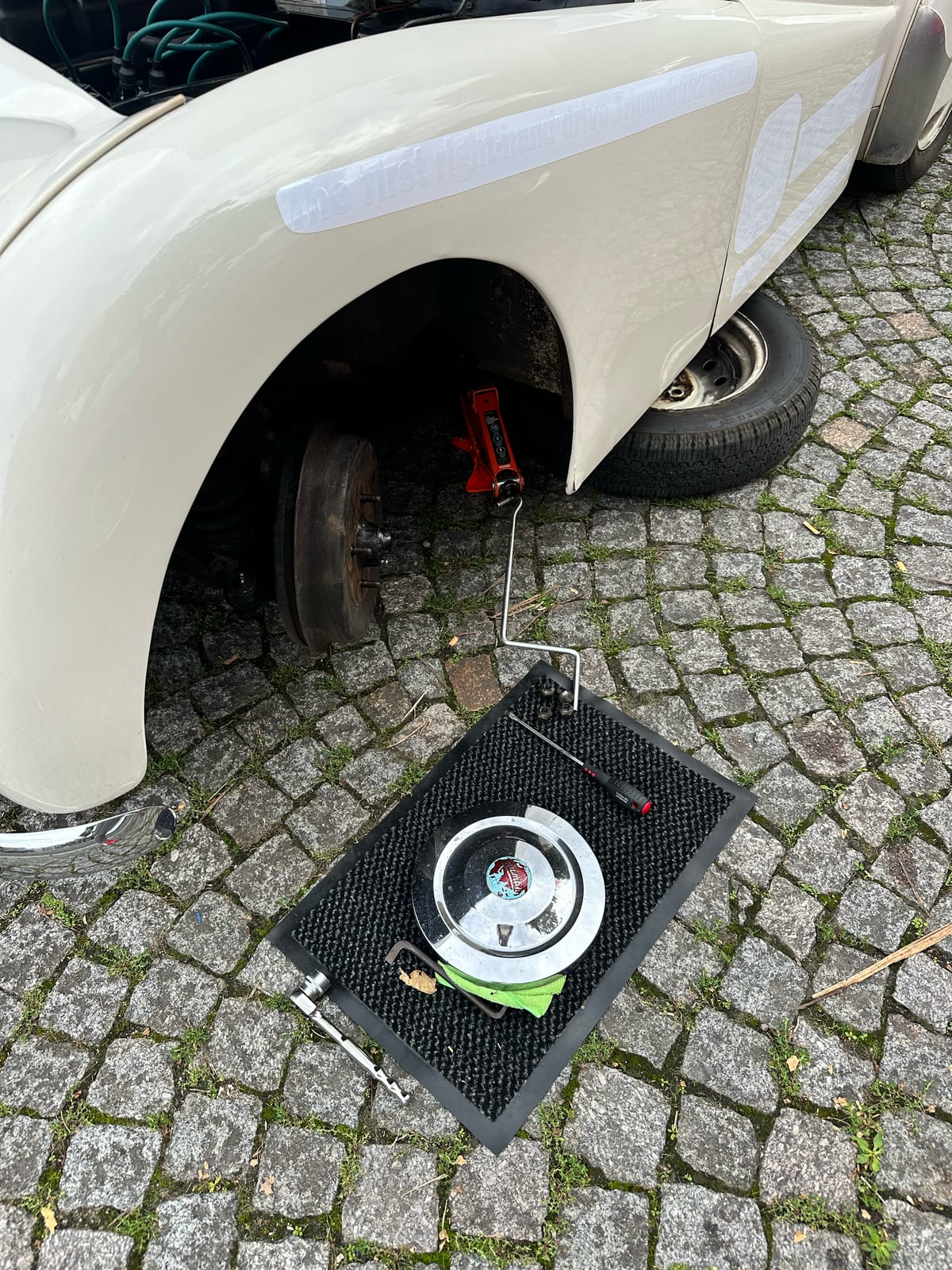 TS2 up on a trolley jack on the cobbles, front wheel off and the bare brake drum showing in the arch, the hubcap and a screwdriver laid out on the doormat