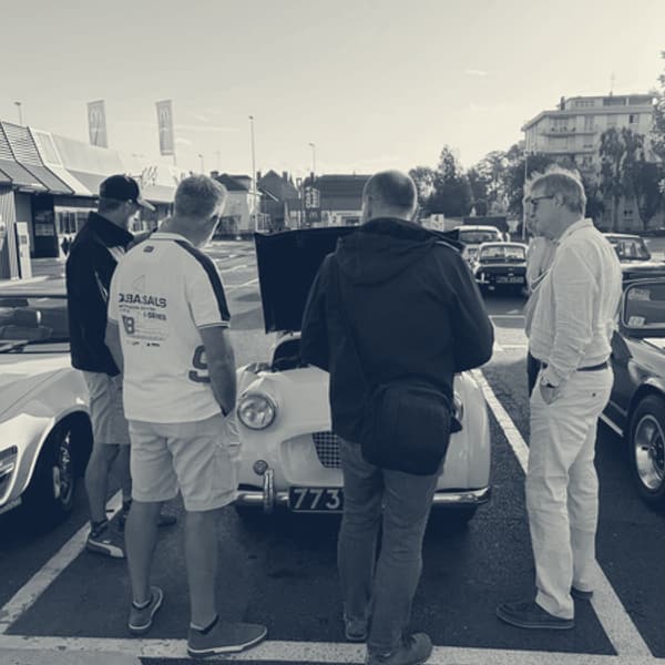 Four men gathered round TS2 with her bonnet up in the supermarket car park, low morning sun behind, a black TR6 in the next bay