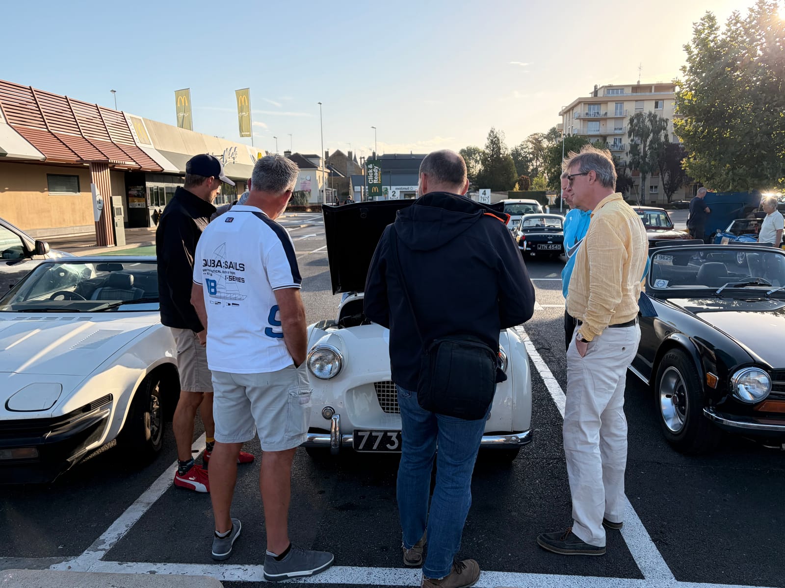 Four men gathered round TS2 with her bonnet up in the supermarket car park, low morning sun behind, a black TR6 in the next bay