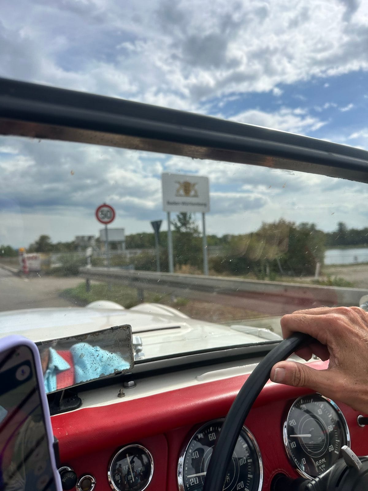 The Baden-Wurttemberg boundary sign with its three lions, seen through TS2's windscreen with a hand on the wheel and the dials below