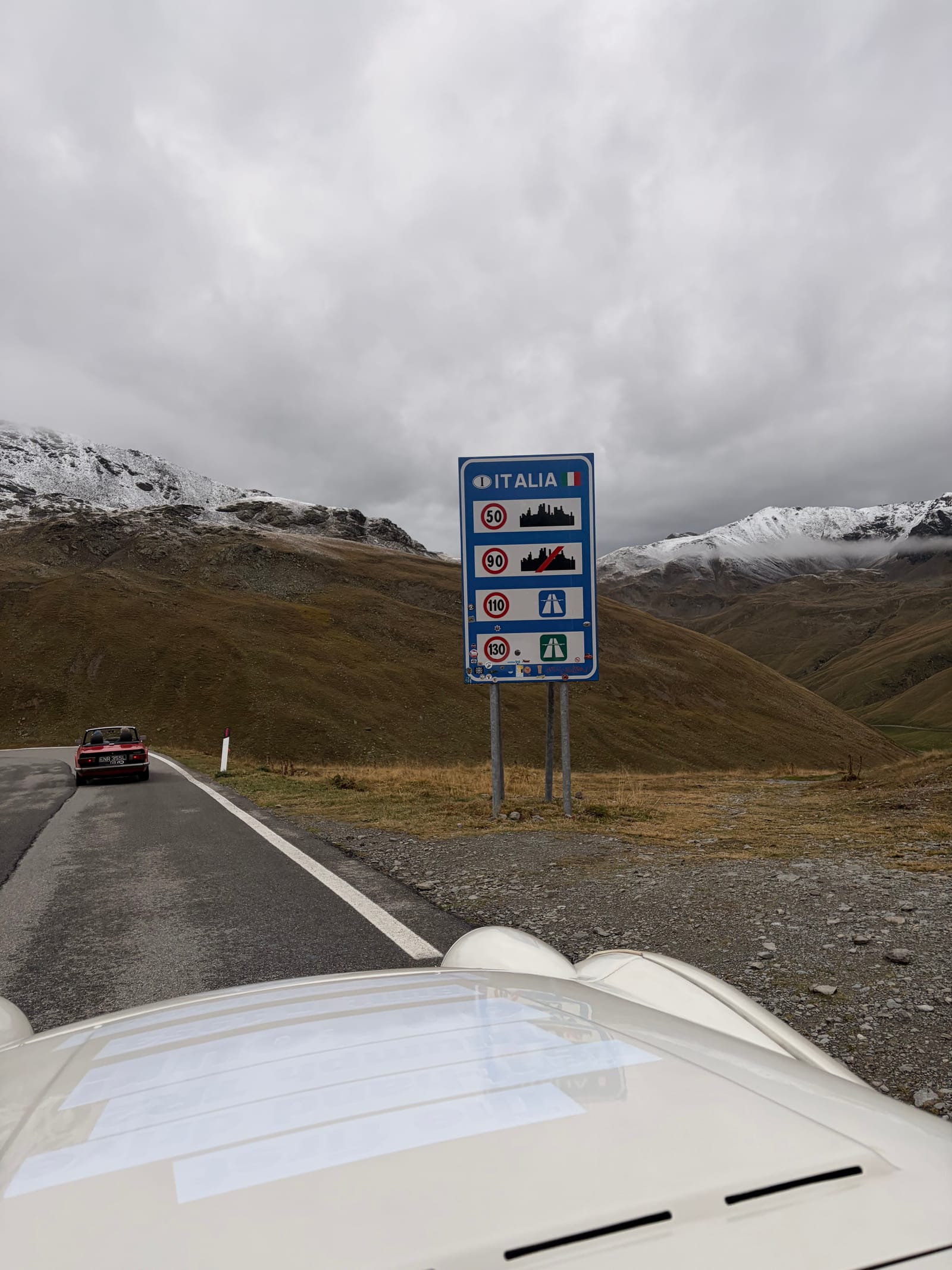 The blue Italia border sign at the top of the Umbrail Pass, seen over TS2's bonnet, a car disappearing down the far side